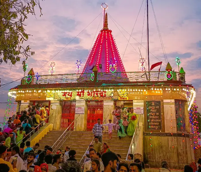 Maihar Maa Sharda Devi Temple beautifully illuminated with colorful lights, as devotees gather on the steps during evening prayers under a scenic sunset sky.