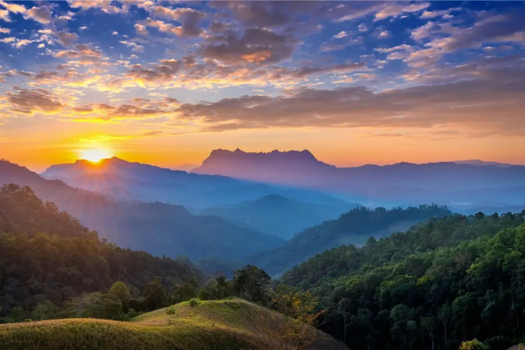 Scenic panoramic view of rolling green hills and mountains at sunrise, with golden sunlight casting a warm glow over the misty landscape and a colorful sky filled with soft clouds.